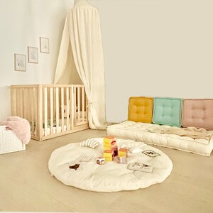 K&ouml;nnte beinhalten: Ein wei&szlig;es Baldachin-Babybett mit einer wei&szlig;en und beigefarbenen Spielmatte auf dem Boden. Die Spielmatte ist mit verschiedenen Spielzeugen verziert, darunter ein Stapel bunter Bl&ouml;cke. Im Hintergrund ist eine hellbraune Couch mit drei Kissen zu sehen. Die Kissen sind gelb, gr&uuml;n und rosa.
