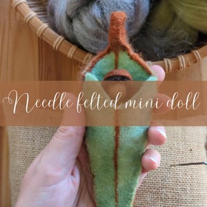May include: A needle felted mini doll in shades of green, brown, and tan. The doll is held in a person's hand. The text "Needle felted mini doll" is overlaid on the image. A basket of wool is in the background.