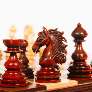 May include: A close-up of a wooden chess set with a carved knight piece in the foreground. The knight is made of dark red wood and has a detailed horse head design. The other pieces are also made of dark red wood and have a polished finish.