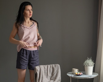 Puede incluir: Una mujer lleva un top de tirantes de lino rosa con un escote de volantes y pantalones cortos de lino gris oscuro. Está de pie sobre una alfombra blanca y esponjosa frente a una pared gris. Hay una silla con una manta marrón y blanca drapeada sobre ella y una mesa pequeña con una taza y un platillo y un plato de caramelos de colores.