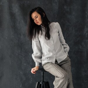 May include: A woman wearing a light gray linen shirt with a white collar and long sleeves, paired with striped gray pants. She is sitting on a black metal stool with her feet resting on the floor.