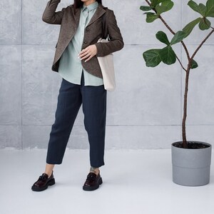 Pode incluir: Uma mulher vestindo um blazer marrom de tweed, uma camisa azul claro de botões, calças azul escuro e um chapéu preto. Ela está segurando uma bolsa tote bege e está em pé em frente a uma parede cinza com uma planta em vaso.