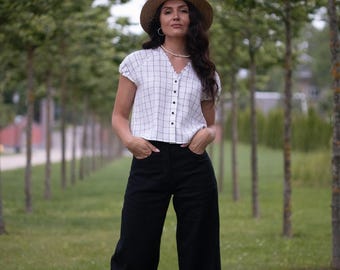 May include: A woman wearing a straw hat, a white plaid top with black buttons, and black wide-leg pants. The outfit is set against a backdrop of green trees and grass, suggesting an outdoor setting.