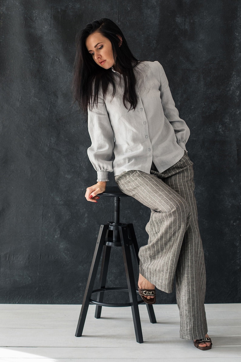 May include: A woman wearing a light gray linen shirt with a white collar and long sleeves, paired with striped gray pants. She is sitting on a black metal stool with her feet resting on the floor.