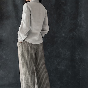 May include: A woman wearing a light gray linen blouse with a ruffled collar and wide-legged striped pants.