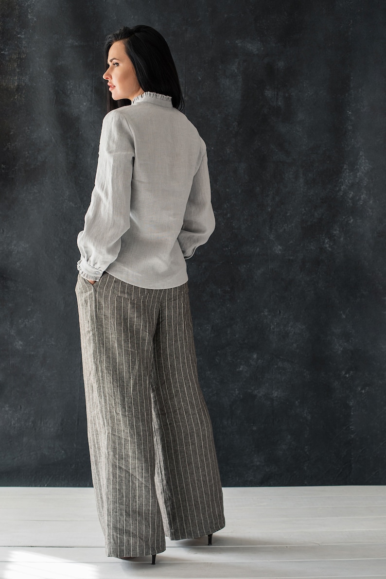 May include: A woman wearing a light gray linen blouse with a ruffled collar and wide-legged striped pants.