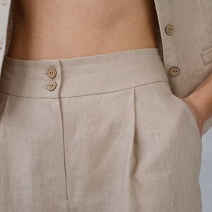 Pode incluir: Cal&ccedil;a de linho bege de cintura alta com pregas. A cal&ccedil;a tem fecho de bot&atilde;o e dois bolsos.