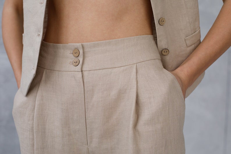 Pode incluir: Cal&ccedil;a de linho bege de cintura alta com pregas. A cal&ccedil;a tem fecho de bot&atilde;o e dois bolsos.