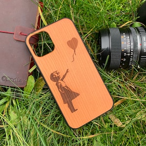 May include: A wooden phone case with a laser-engraved design of a girl reaching up to a heart-shaped balloon. The case is made of natural wood with a light brown color.