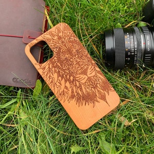 May include: A wooden phone case featuring a detailed laser-engraved design of a lion's face. The case is light brown and the design is dark brown. The case is lying on a bed of green grass.