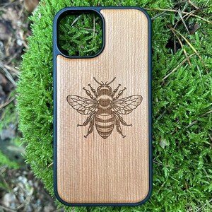 Könnte beinhalten: Eine Handyhülle aus Holz mit schwarzem Rand. Die Hülle zeigt eine detaillierte Gravur einer Biene in der Mitte. Das Holz hat eine hellbraune Farbe und ein sichtbares Maserungsmuster. Die Hülle liegt auf grünem Moos.