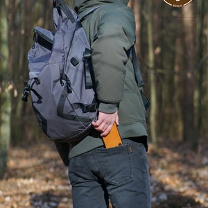 May include: A person wearing a green hooded jacket and blue jeans is holding a wooden folding chair in their back pocket. The chair is closed and has a brown leather strap. The person is wearing a gray backpack. The background is a forest.