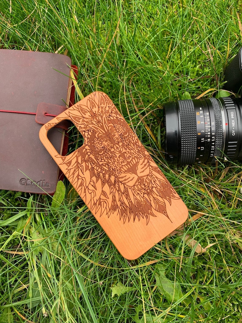 K&ouml;nnte beinhalten: Eine Handyh&uuml;lle aus Holz mit einem lasergravierten Design eines L&ouml;wenkopfes. Das Design ist filigran und detailliert, mit einem mandalaartigen Muster.