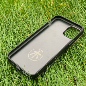 May include: Black phone case with a gold trim and a gold logo that says "atelier" with a stylized tree symbol. The case is lying on green grass.