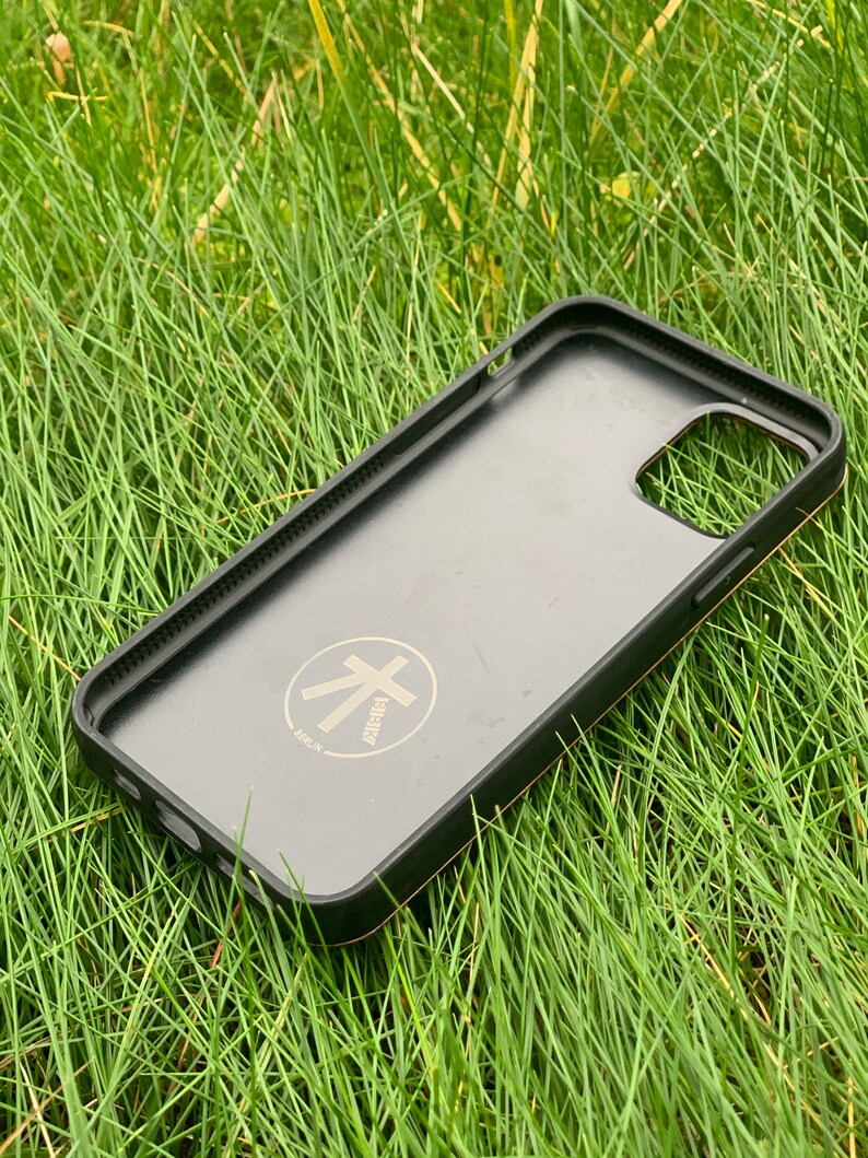 May include: Black phone case with a gold trim and a gold logo that says "atelier" with a stylized tree symbol. The case is lying on green grass.