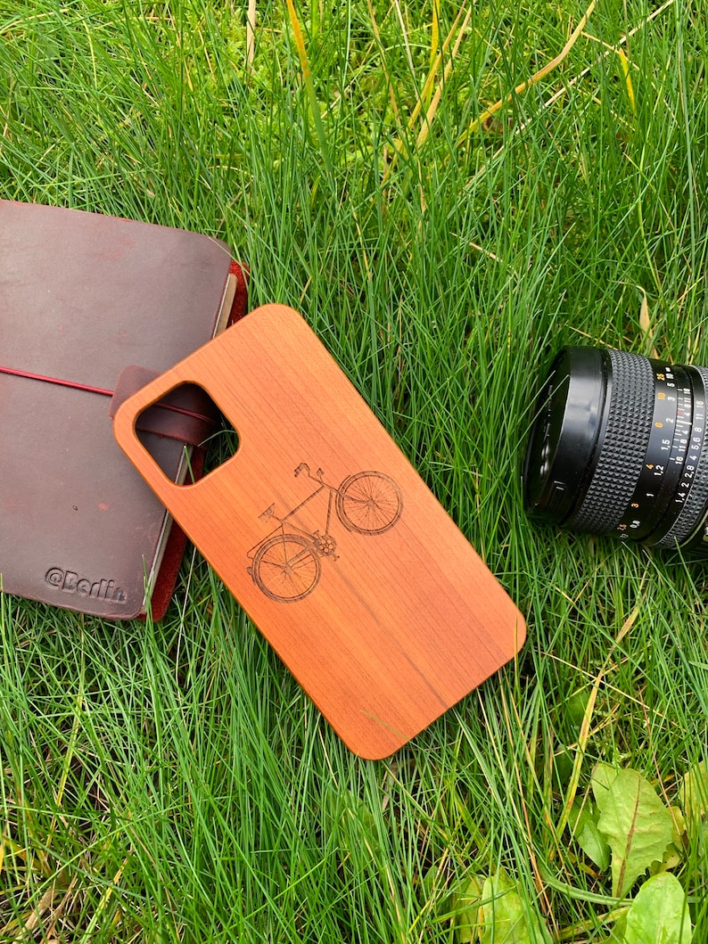 May include: A wooden phone case with a laser-engraved image of a bicycle. The case is a light brown color and has a natural wood grain. The phone case is lying on a bed of green grass.