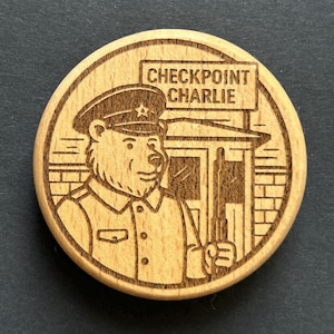 Könnte beinhalten: Ein runder Holz-Souvenir mit einem geätzten Design. Es zeigt einen Bären in Militäruniform mit einem Gewehr vor einem Kontrollpunkt. Der Text "CHECKPOINT CHARLIE" steht darüber. Das Design ist im Vintage-Stil mit aufwendigen Details.