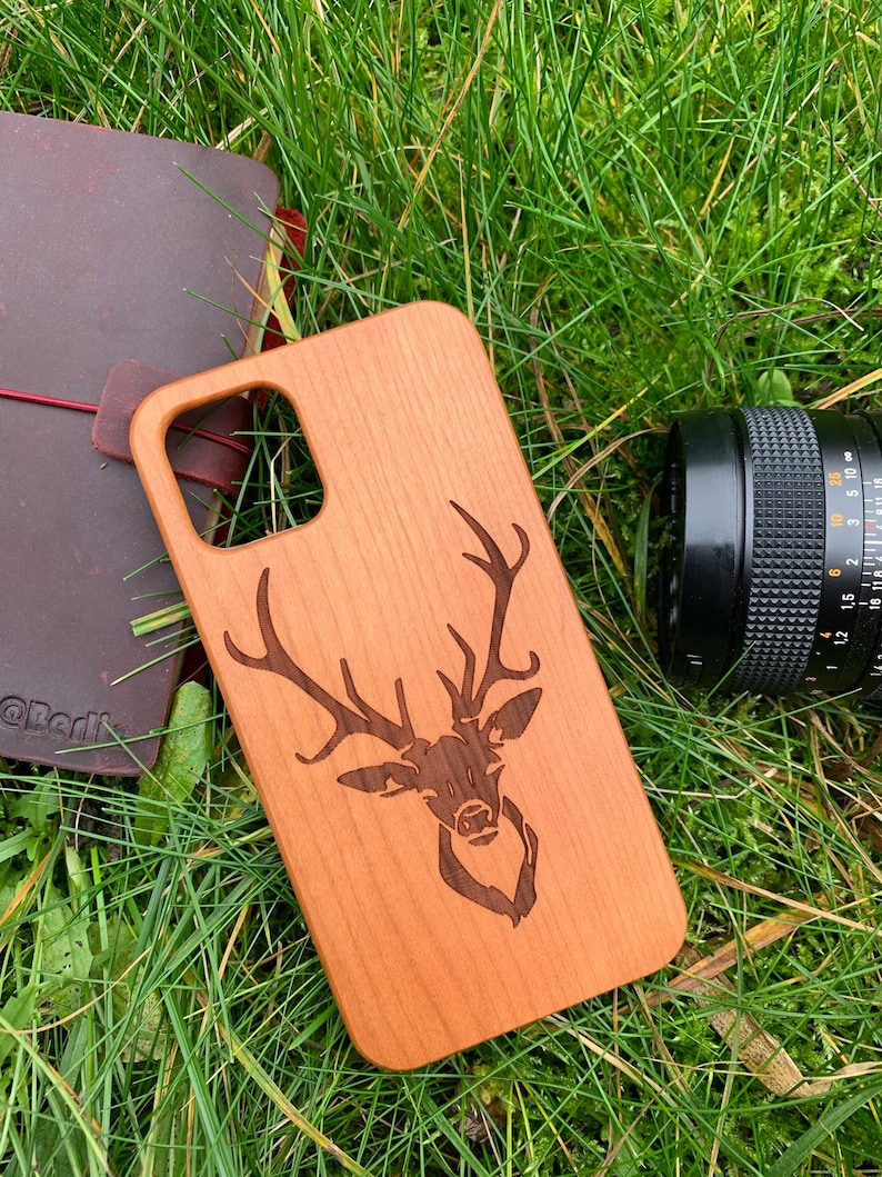 May include: A wooden phone case with a laser-engraved design of a deer head with antlers. The case is a light brown color and has a natural wood grain.