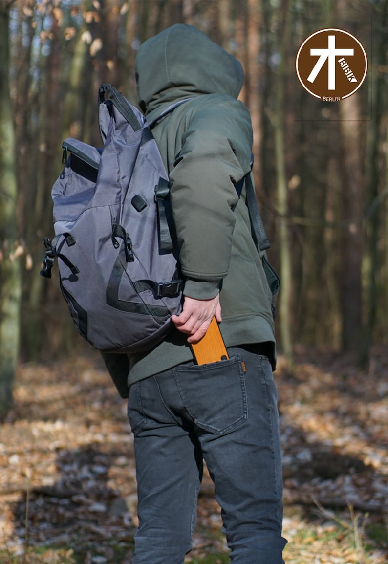 K&ouml;nnte beinhalten: Eine Person tr&auml;gt eine gr&uuml;ne Kapuzenjacke und eine blaue Jeans und h&auml;lt einen Holz-Klappstuhl in der Ges&auml;&szlig;tasche. Der Stuhl ist zusammengeklappt und hat einen braunen Lederriemen. Die Person tr&auml;gt einen grauen Rucksack. Der Hintergrund ist ein Wald.