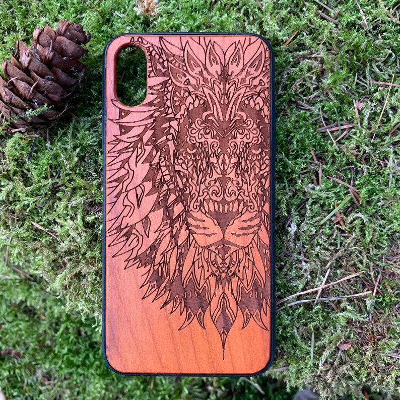 K&ouml;nnte beinhalten: Eine Handyh&uuml;lle aus Holz mit einem lasergravierten Design eines L&ouml;wenkopfes. Das Design ist filigran und detailliert, mit einem mandalaartigen Muster um den L&ouml;wenkopf herum. Die H&uuml;lle ist in einem satten Rotbraun gehalten.