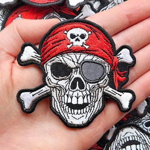 May include: Embroidered pirate skull patch with a red bandana, eye patch, and crossed bones. The skull is white with black details, and the bandana features a small skull emblem. A collection of similar patches are in the background.