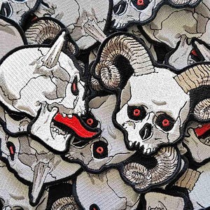 May include: A collection of embroidered patches featuring skull designs. The skulls are primarily white with gray shading, red eyes, and some have horns or open mouths with red tongues. The patches are densely packed together.