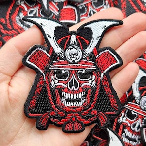 May include: Embroidered patch featuring a red and black samurai skull design. The skull wears a helmet with white accents. The patch is held in a hand, with other similar patches in the background. The patches are likely for clothing or accessories.