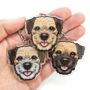 May include: Three dog face keychains with a clear background. Each keychain features a detailed illustration of a dog's face, with varying shades of brown, tan, and black fur. The keychains are held together by a silver chain.
