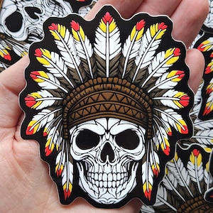 May include: A sticker depicting a skull adorned with a Native American headdress. The skull is white with black eye sockets and a detailed jawline. The headdress is brown with white, yellow, and red feathers. The sticker has a black border.