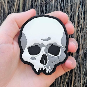 May include: A skull-shaped patch with a black border. The skull is white and grey, with black eye sockets and a nose cavity. The patch is held in a hand against a background of brown twigs.
