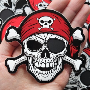 May include: A pirate skull sticker featuring a red bandana, black eye patch, and crossed bones. The skull is white with black detailing, and the bandana has a white skull and crossbones. The sticker is held in a hand, surrounded by similar stickers.