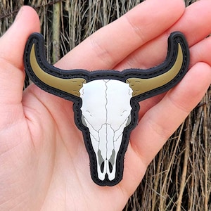 May include: A black, tan, and white patch in the shape of a bull skull. The skull is white with tan horns and a black border. The patch is held in a hand against a background of brown twigs.