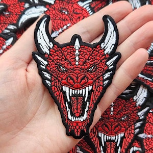 May include: A red dragon patch with white horns and teeth, and a black border. The patch is held in a hand, with many similar patches in the background. The dragon has an open mouth with visible teeth.