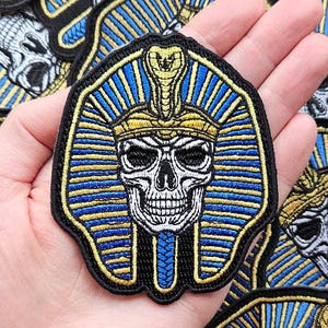 May include: Embroidered patch featuring a skull wearing a golden pharaoh headdress with a cobra. The patch has a blue and gold color scheme with a black border. The skull has a detailed design.