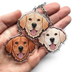 May include: Three golden retriever dog charms on a silver chain keyring. The charms are made of clear acrylic and feature hand-painted portraits of the dogs. The dogs are all smiling with their tongues out.