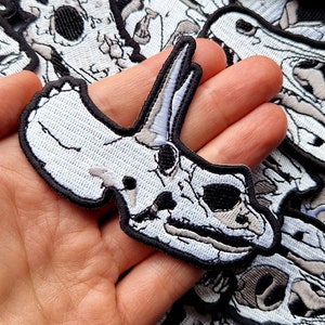 May include: A white embroidered patch of a dinosaur skull with black detailing. The patch is held in a hand.