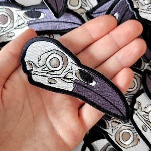 Peut inclure: Écusson brodé représentant un crâne de corbeau. Le patch présente un crâne blanc avec des détails gris et noirs, et un bec violet. Il a une bordure noire. Le fond montre d'autres écussons du même motif.
