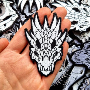 May include: White embroidered iron-on patch of a dragon skull with a detailed design. The patch is held in a hand.
