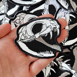Peut inclure: Un gros plan sur un patch brodé de crâne de serpent blanc sur un fond de tissu noir. Le patch est tenu dans une main. Le patch est entouré d'autres patchs de crâne de serpent.