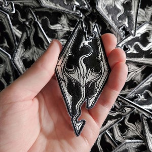 Peut inclure: Un patch brodé noir et blanc représentant le symbole du dragon du jeu vidéo The Elder Scrolls V: Skyrim. Le patch est tenu dans une main.