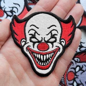 May include: An embroidered patch featuring a clown face with white skin, a bright red nose, and red hair. The clown has a wide, menacing grin with sharp, pointed teeth and black detailing around the eyes and mouth, all outlined in a thick black border.