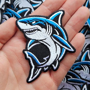 May include: An embroidered patch featuring a shark with white, light blue, and dark blue thread detailing. The shark is depicted in a dynamic swimming pose with its mouth slightly open, showing teeth, and is outlined with a thick black border.