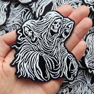 May include: An embroidered patch featuring a black and white design of a three-headed skeletal figure draped in flowing robes. The intricate stitching highlights the skulls and detailed fabric folds against a solid black background.