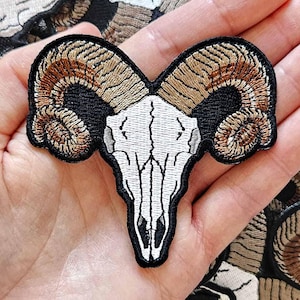 Peut inclure: Écusson brodé représentant un crâne de bélier avec de grandes cornes incurvées. L'écusson a une bordure noire, avec le crâne en blanc et les cornes dans des tons bruns. L'écusson est tenu dans une main, avec d'autres écussons similaires en arrière-plan.