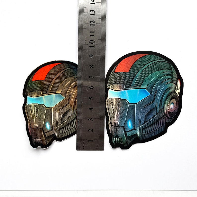 Pode incluir: Dois adesivos de capacete futuristas com uma r&eacute;gua para escala. Os adesivos apresentam um design detalhado com tons de cinza, preto e azul, com um detalhe vermelho no topo. A &aacute;rea da viseira tem um brilho azul brilhante. A r&eacute;gua mostra cerca de 13 cm de altura.