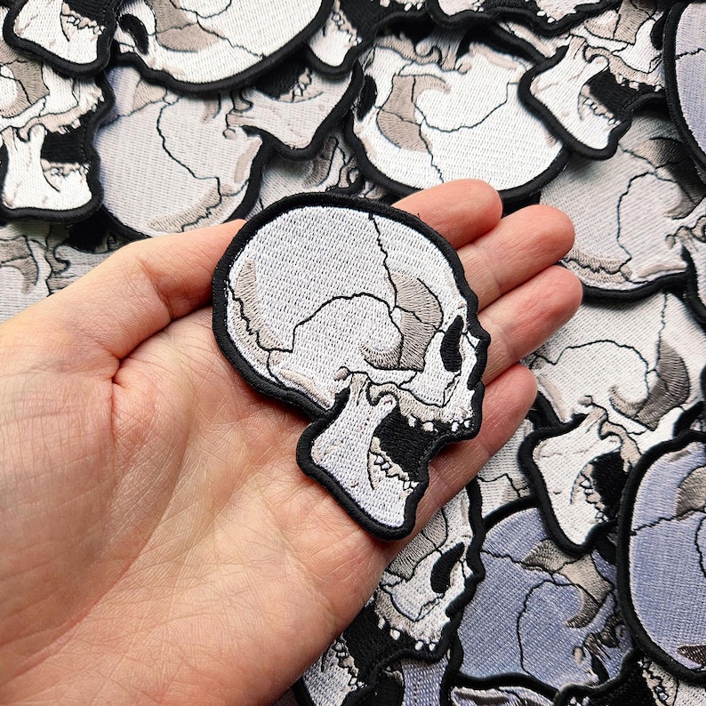 K&ouml;nnte beinhalten: Ein wei&szlig;er gestickter Totenkopf-Patch mit offenem Mund und sichtbaren Z&auml;hnen. Der Patch wird in einer Hand vor einem Hintergrund aus anderen Totenkopf-Patches gehalten.