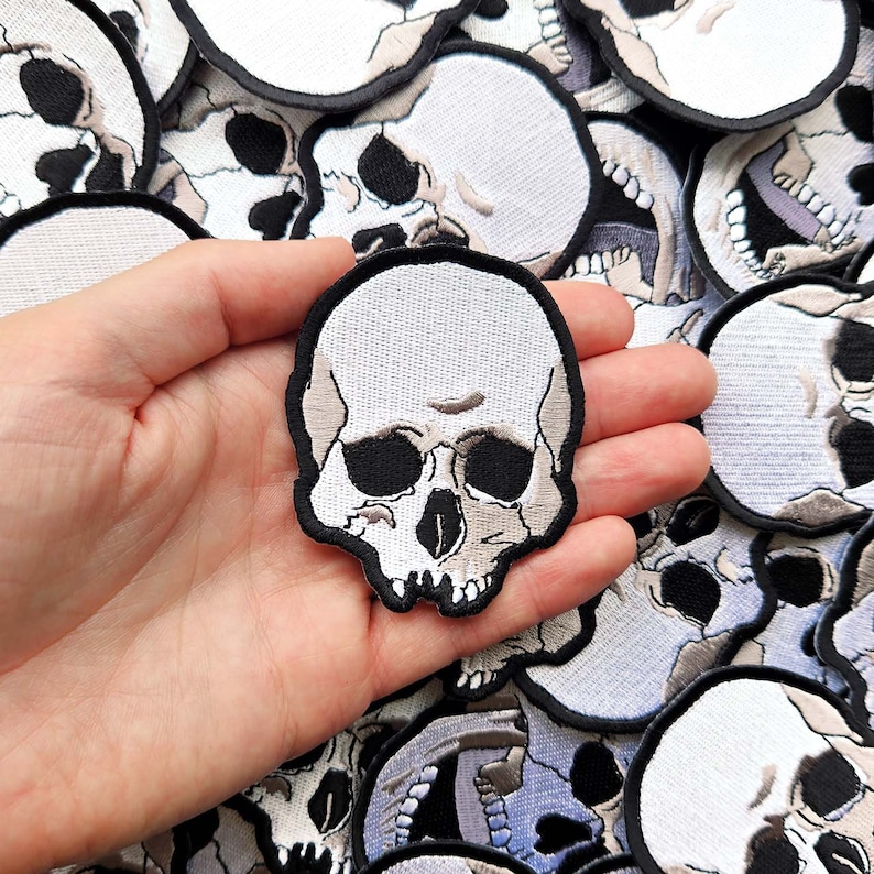 K&ouml;nnte beinhalten: Ein wei&szlig;er gestickter Totenkopf-Patch mit schwarzen Augenh&ouml;hlen und einer detaillierten Kinnlinie. Der Patch wird in einer Hand vor einem Hintergrund aus anderen Totenkopf-Patches gehalten.