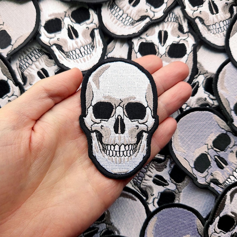 K&ouml;nnte beinhalten: Ein wei&szlig;er gestickter Totenkopf-Patch mit schwarzen Augenh&ouml;hlen und Z&auml;hnen. Der Patch wird in einer Hand vor einem Hintergrund aus vielen anderen Totenkopf-Patches gehalten.
