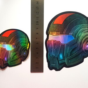 Pode incluir: Dois autocolantes hologr&aacute;ficos com um design de capacete futurista. Os capacetes t&ecirc;m um gradiente de cor vermelho, verde e azul. Uma r&eacute;gua &eacute; colocada ao lado dos autocolantes para compara&ccedil;&atilde;o de tamanho.