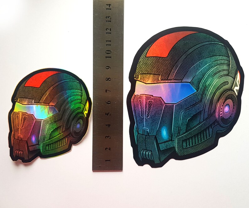 Pode incluir: Dois autocolantes hologr&aacute;ficos com um design de capacete futurista. Os capacetes t&ecirc;m um gradiente de cor vermelho, verde e azul. Uma r&eacute;gua &eacute; colocada ao lado dos autocolantes para compara&ccedil;&atilde;o de tamanho.